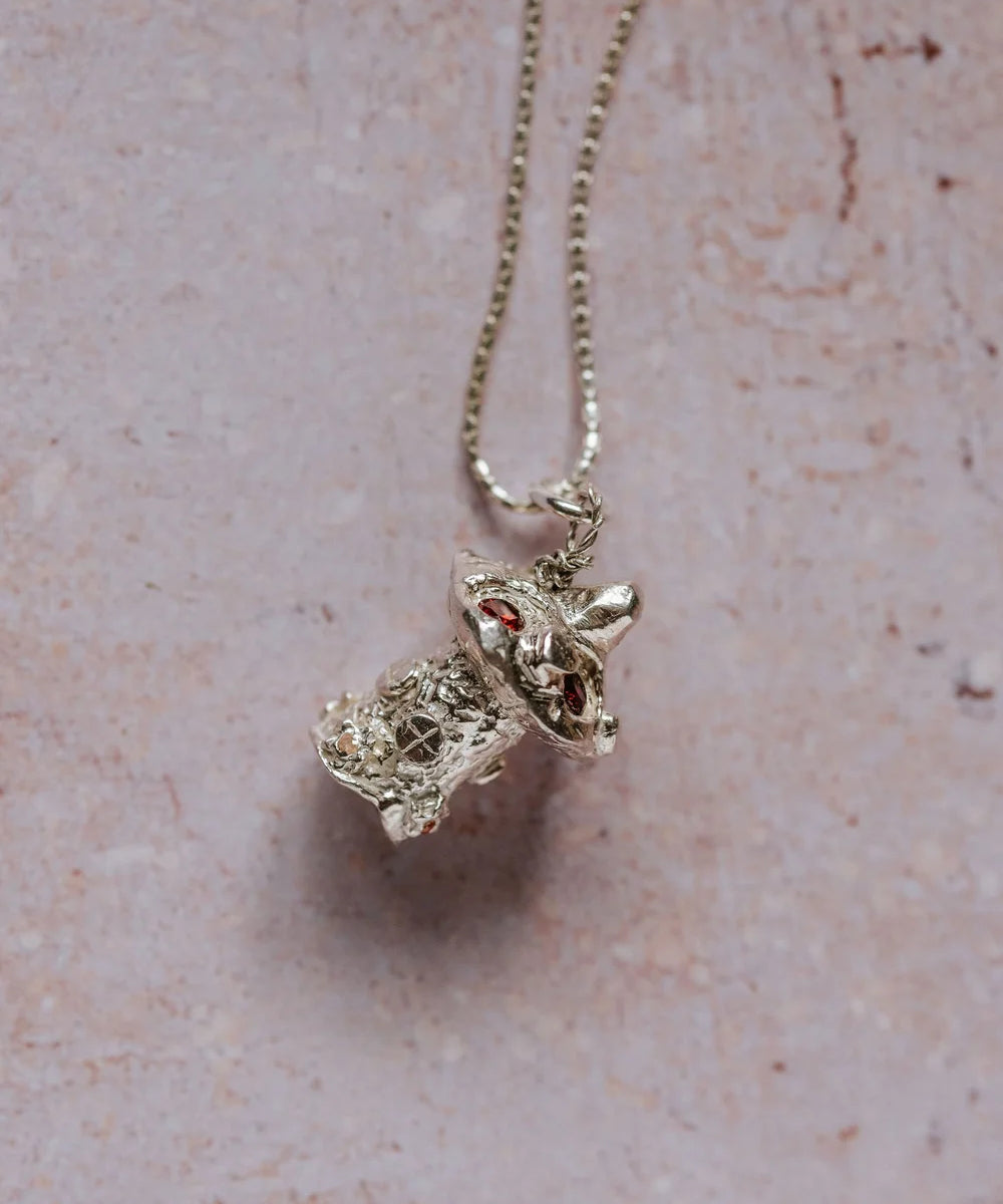 Angled close-up of handmade silver mushroom toadstool necklace with red cubic zirconia stones, displayed on pink background