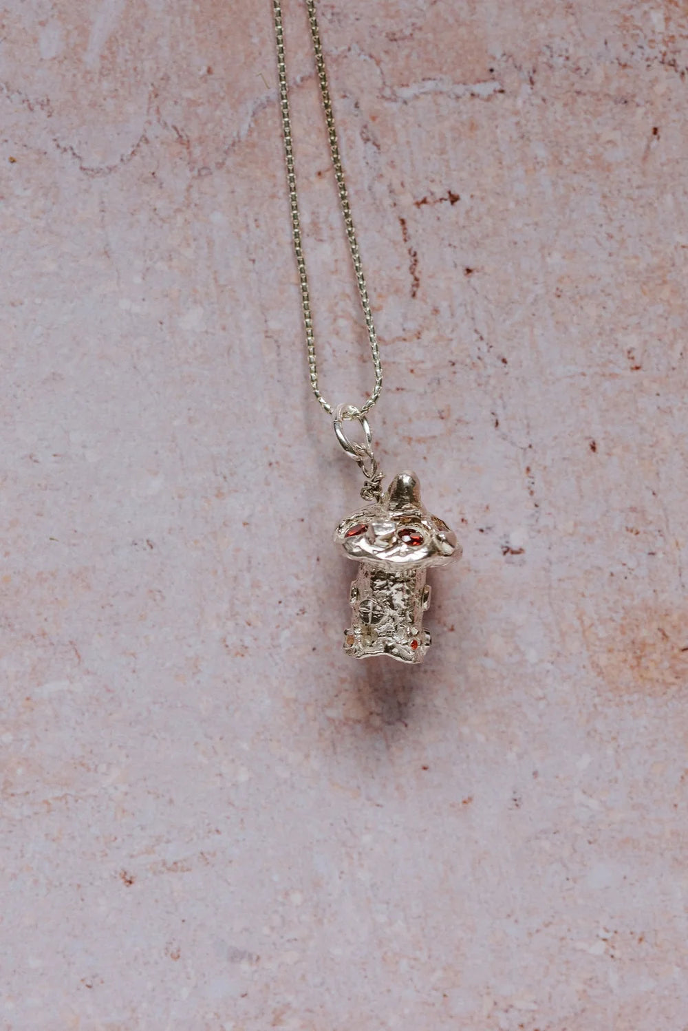 Full view of silver mushroom toadstool pendant necklace with red gemstones, on a 16-inch silver chain against a pink surface