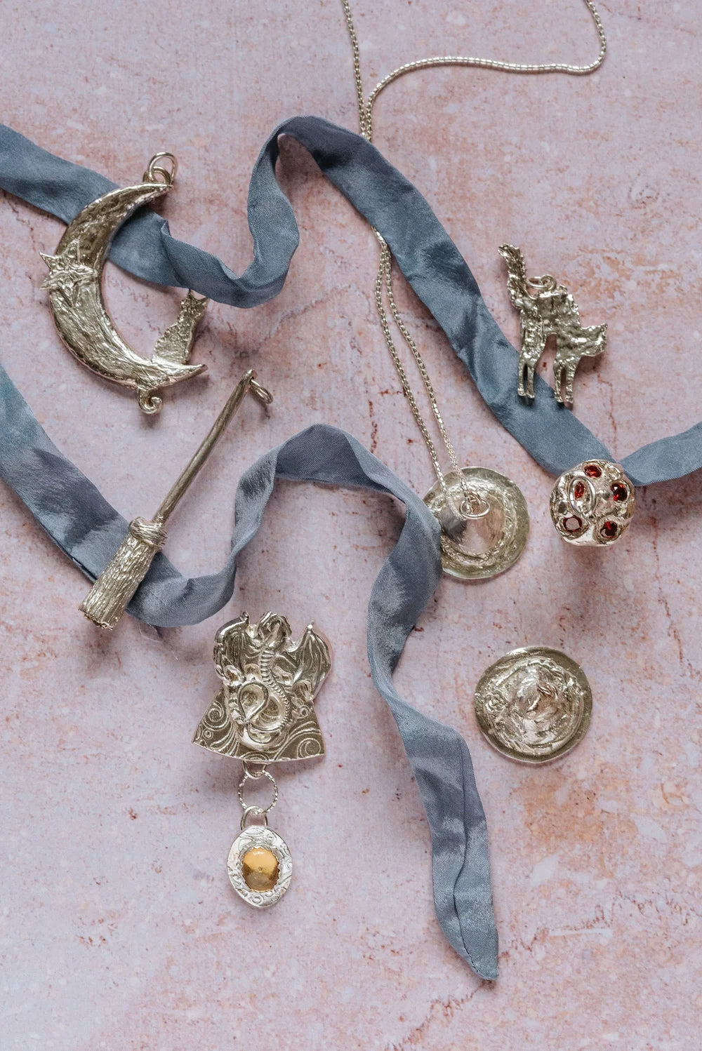 Flatlay of handmade silver mystical jewellery including a mushroom toadstool pendant, crescent moon charm, broomstick, cat, and dragon designs styled with silk ribbon on a soft pink background