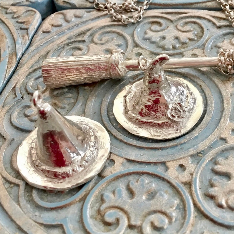 Handmade recycled silver witch hat pendants, one with tilted tip and floral detail on brim, pictured beside matching broomstick pendant