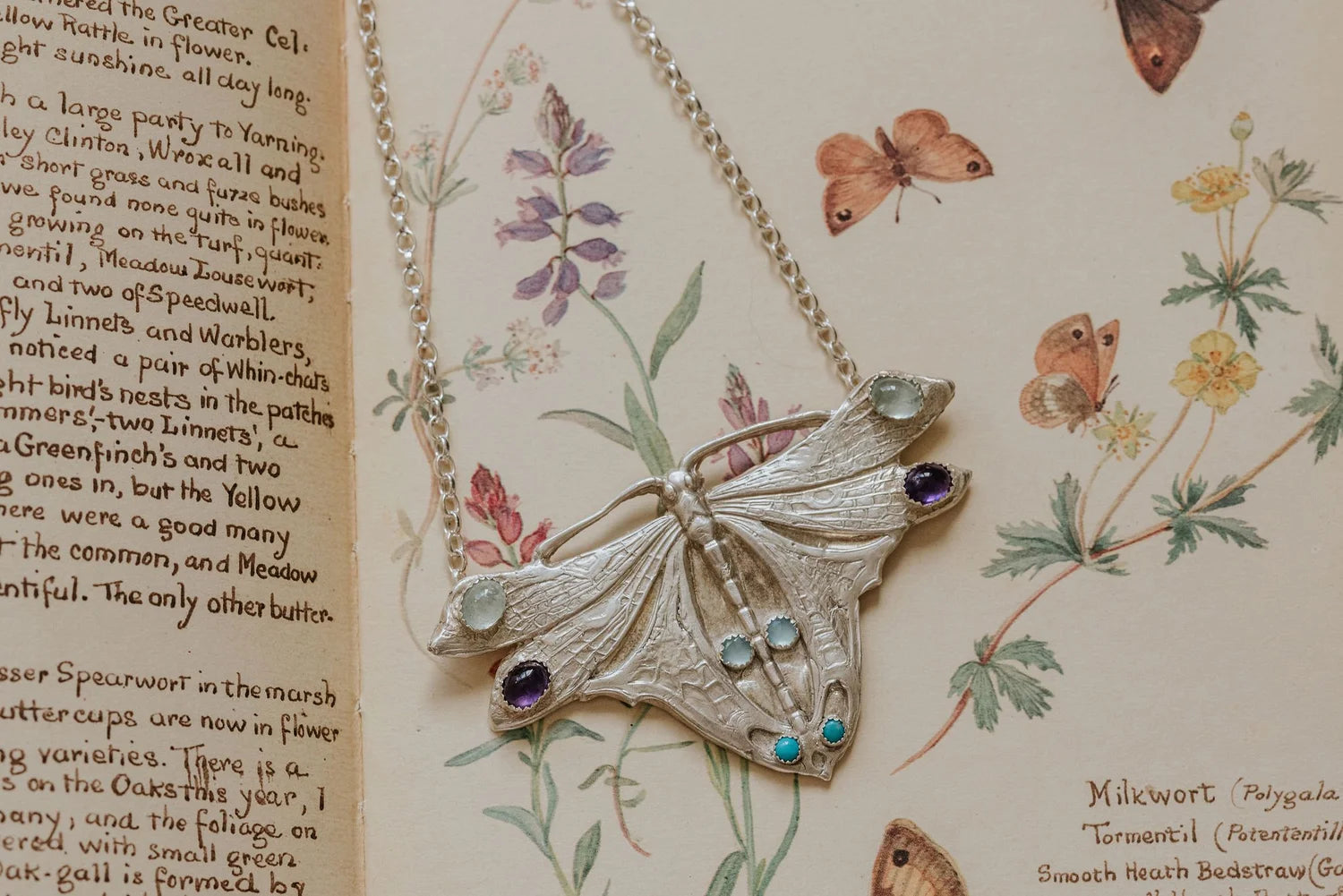 Handmade Art Nouveau inspired silver moth statement necklace with amethyst, turquoise and aquamarine gemstones displayed on vintage floral illustration background.