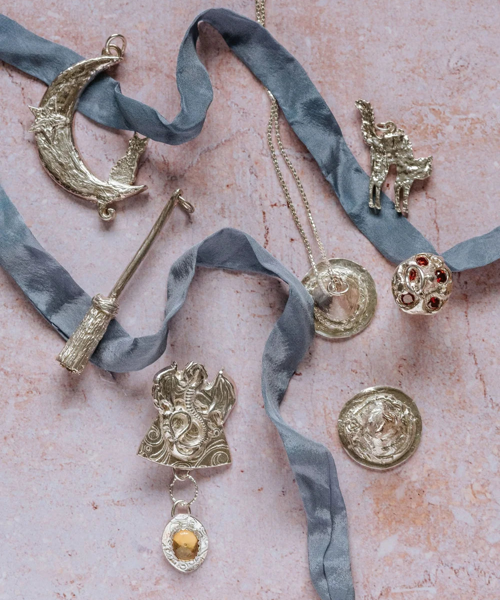 Flat lay of unique handmade silver jewellery including dragon pendant with citrine, crescent moon necklace, broomstick pendant, and whimsical designs from The Magic of Fireflies and Dragonflies