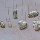 Flatlay of six unique silver leaf necklaces on a blush stone background, highlighting handcrafted texture and fine silver finish.