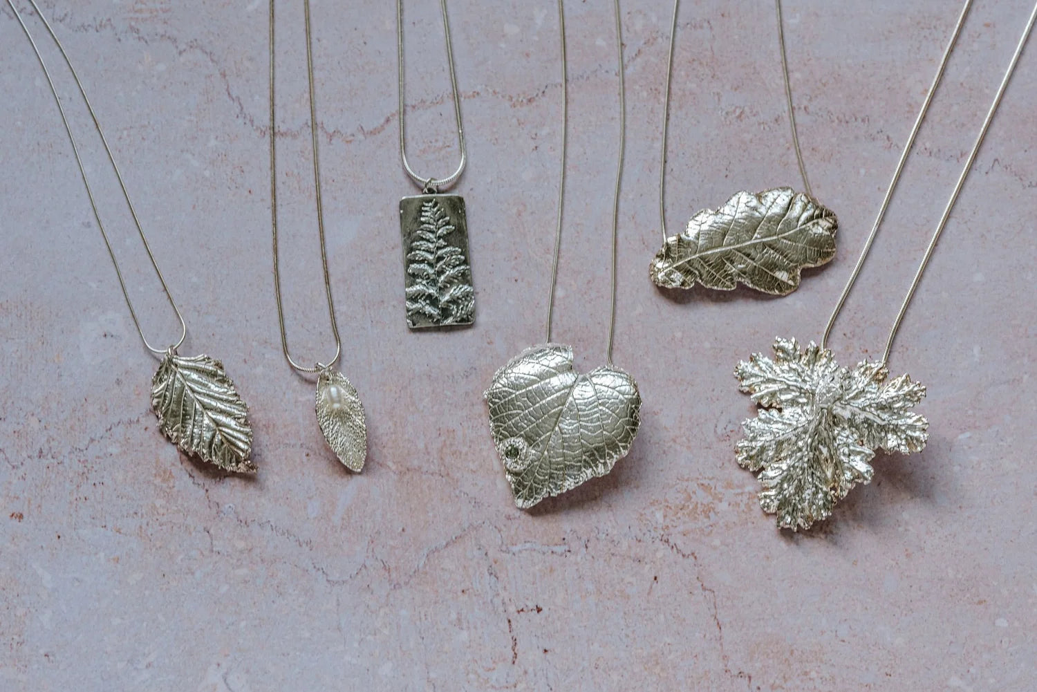 Flatlay of six unique silver leaf necklaces on a blush stone background, highlighting handcrafted texture and fine silver finish.
