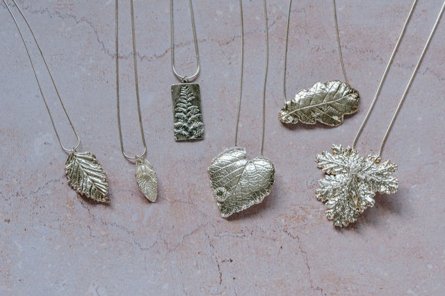 Flatlay of six unique silver leaf necklaces on a blush stone background, highlighting handcrafted texture and fine silver finish.