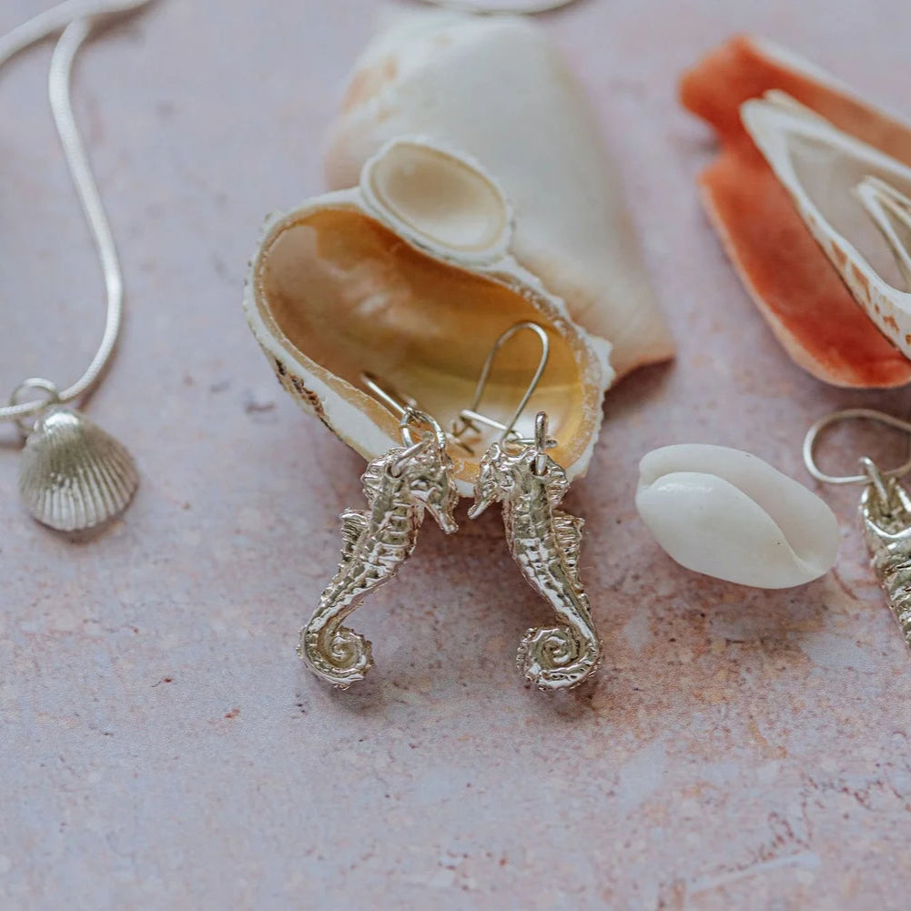 Handmade silver coastal jewellery including seahorse earrings, shell pendant necklaces, and ocean-inspired charms displayed with seashells.