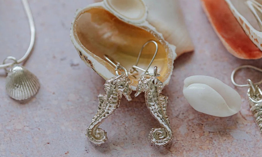 Handmade silver coastal jewellery including seahorse earrings, shell pendant necklaces, and ocean-inspired charms displayed with seashells.