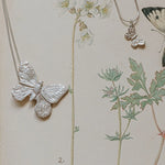 Large and small recycled silver butterfly pendants on snake chains, displayed on botanical illustration background