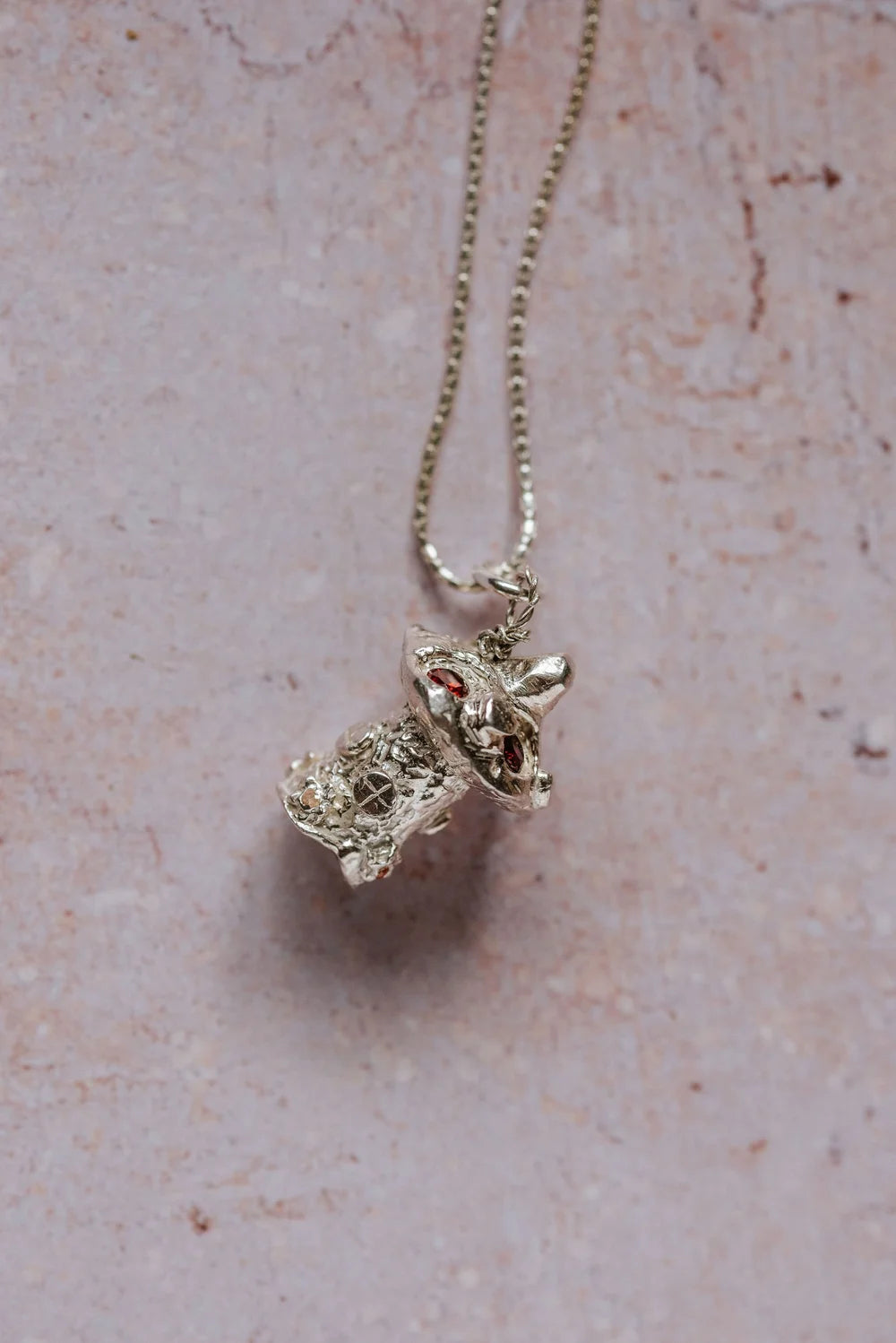 Angled close-up of handmade silver mushroom toadstool necklace with red cubic zirconia stones, displayed on pink background

