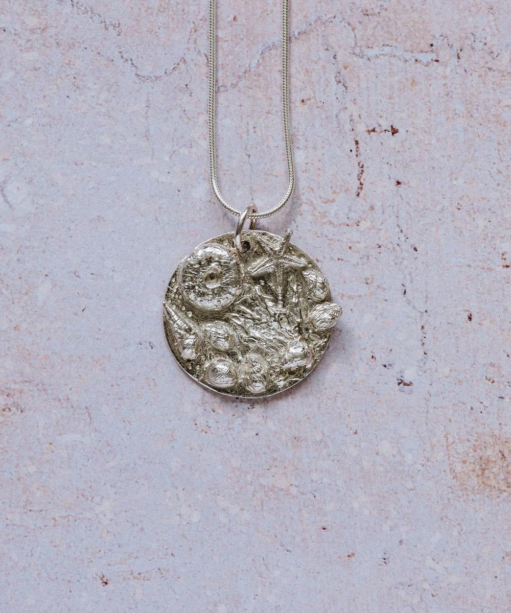 Handmade recycled silver rockpool pendant with starfish and shells on 16-inch chain, displayed on textured stone background