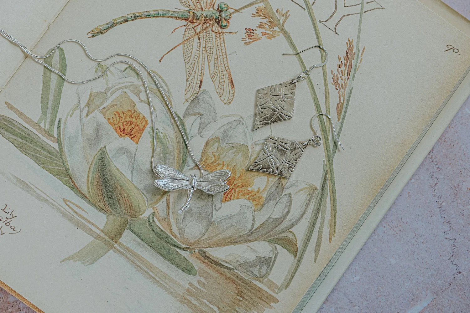 a pair of silver dragonfly earrings with a silver dragonfly necklace on an open botanical illustrated book