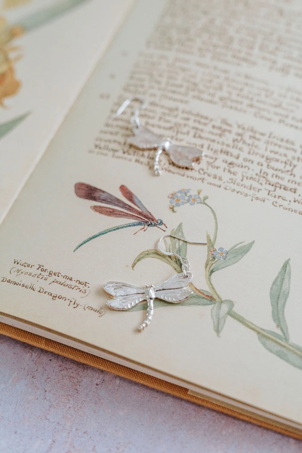 Handmade silver dragonfly earrings displayed on vintage botanical book illustration with forget-me-not flowers.