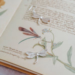 Handmade silver dragonfly earrings displayed on vintage botanical book illustration with forget-me-not flowers.
