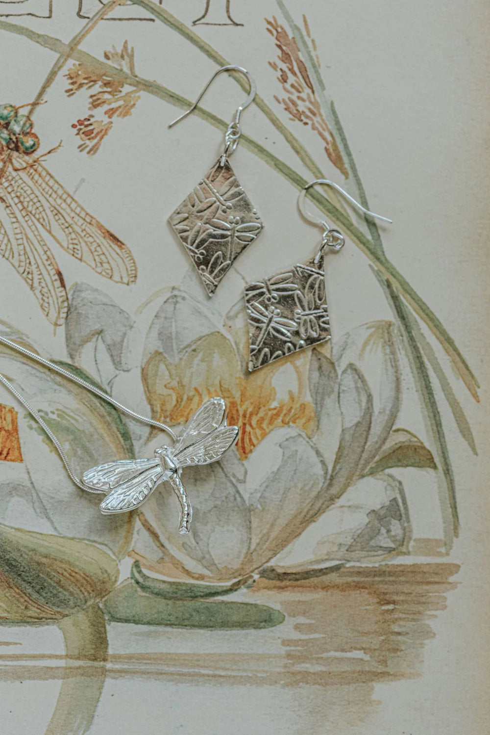 Silver dragonfly earrings with intricate designs on a botanical illustration background