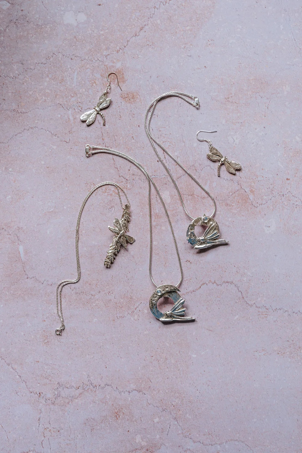 Flat lay of handmade silver dragonfly jewellery collection on pink background, including dragonfly earrings, moon dragonfly pendants, and catkin dragonfly pendant, artisan nature inspired jewellery gifts UK