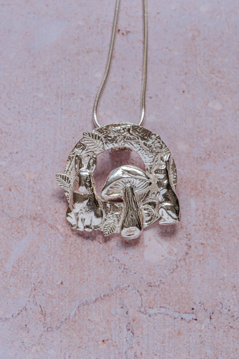 Handmade silver woodland pendant necklace featuring two hares and a mushroom framed by textured leaves, crafted from fine silver on a chain.