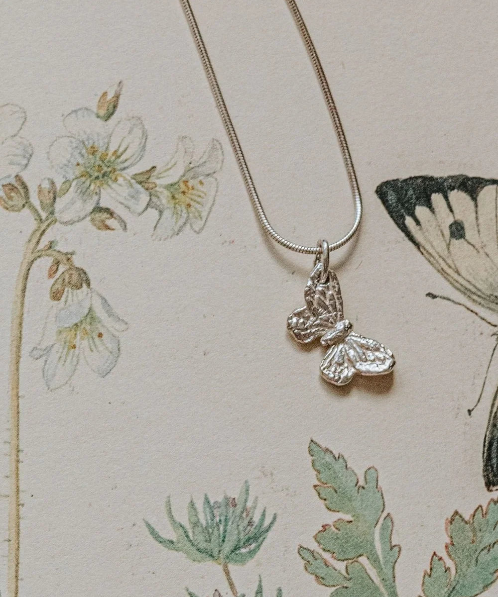Handmade recycled silver small butterfly pendant on 18-inch snake chain, displayed on botanical illustration background