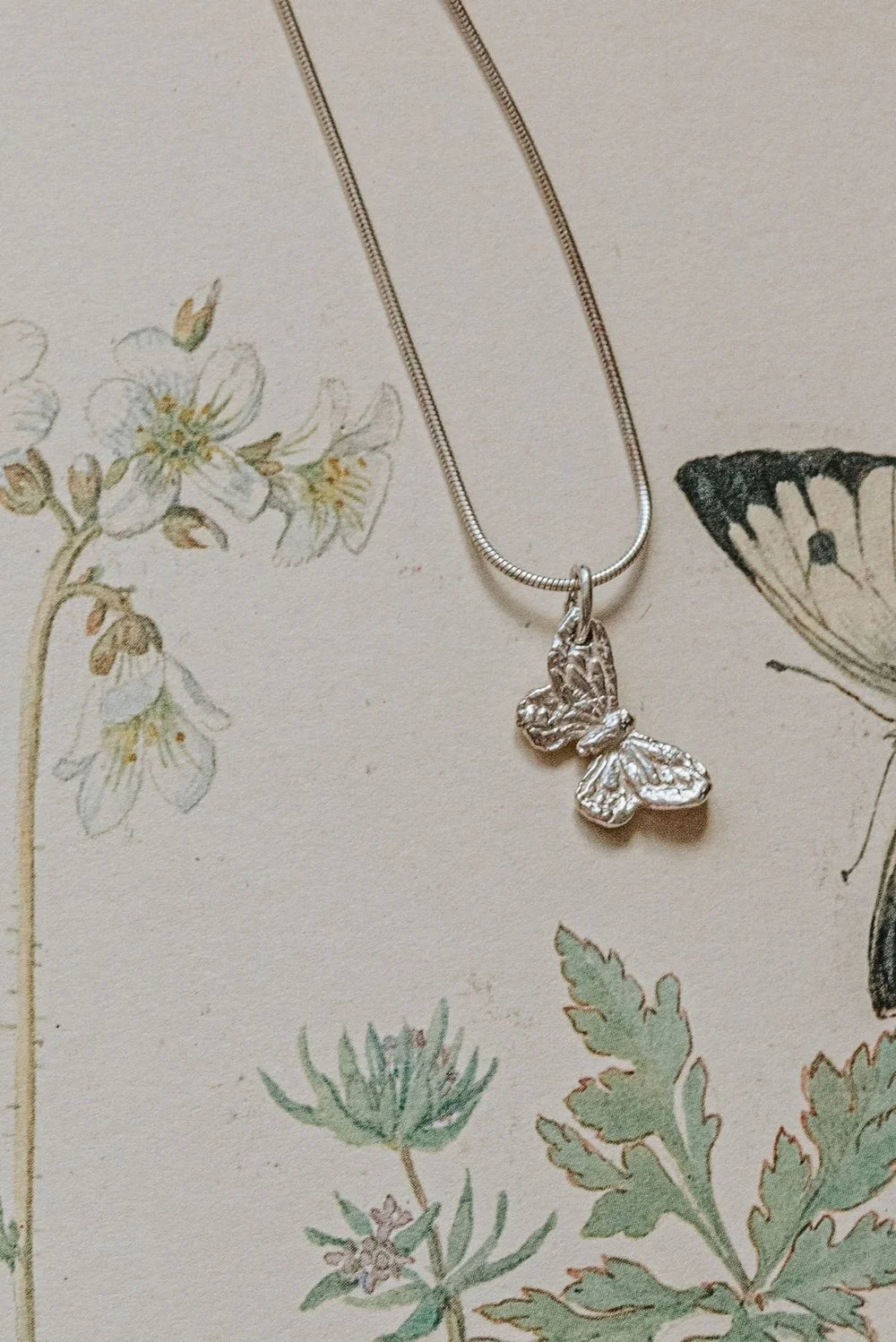 Handmade recycled silver small butterfly pendant on 18-inch snake chain, displayed on botanical illustration background