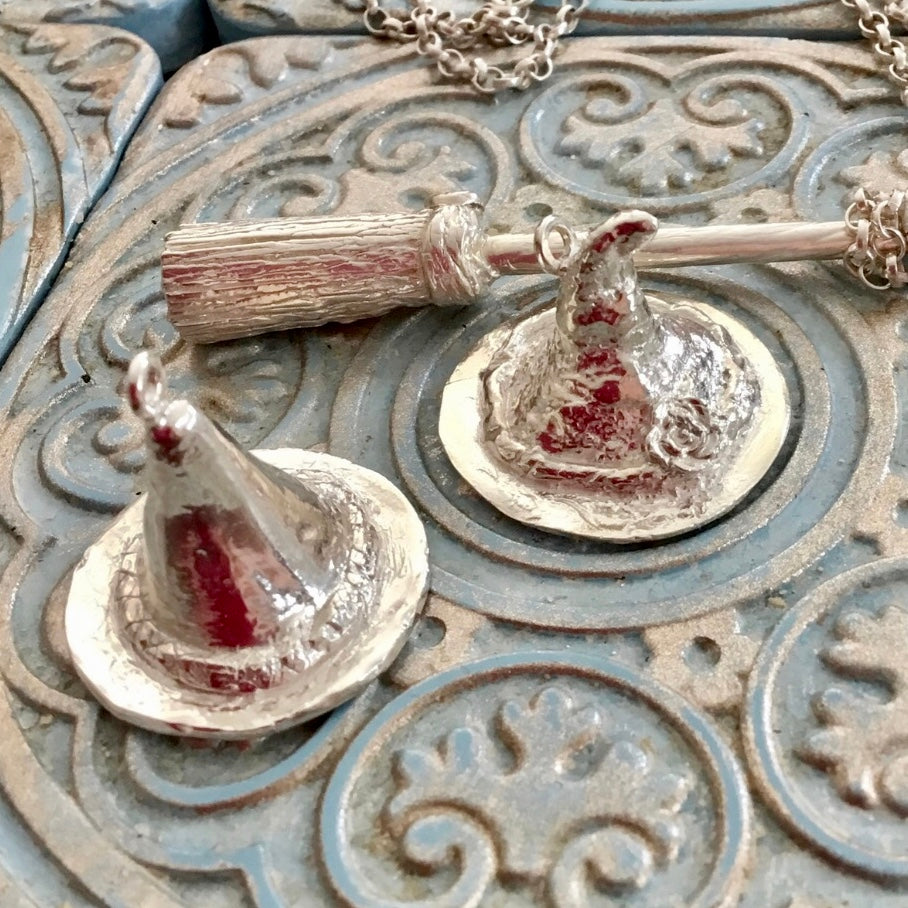 Handmade recycled silver witch hat pendants, one with tilted tip and floral detail on brim, pictured beside matching broomstick pendant